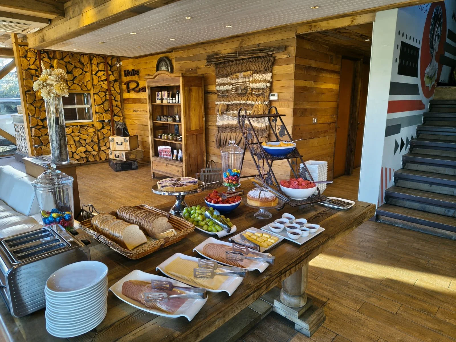 Desayuno estilo buffet con variedad de panes, frutas, cereales y dulces en un ambiente rústico de madera