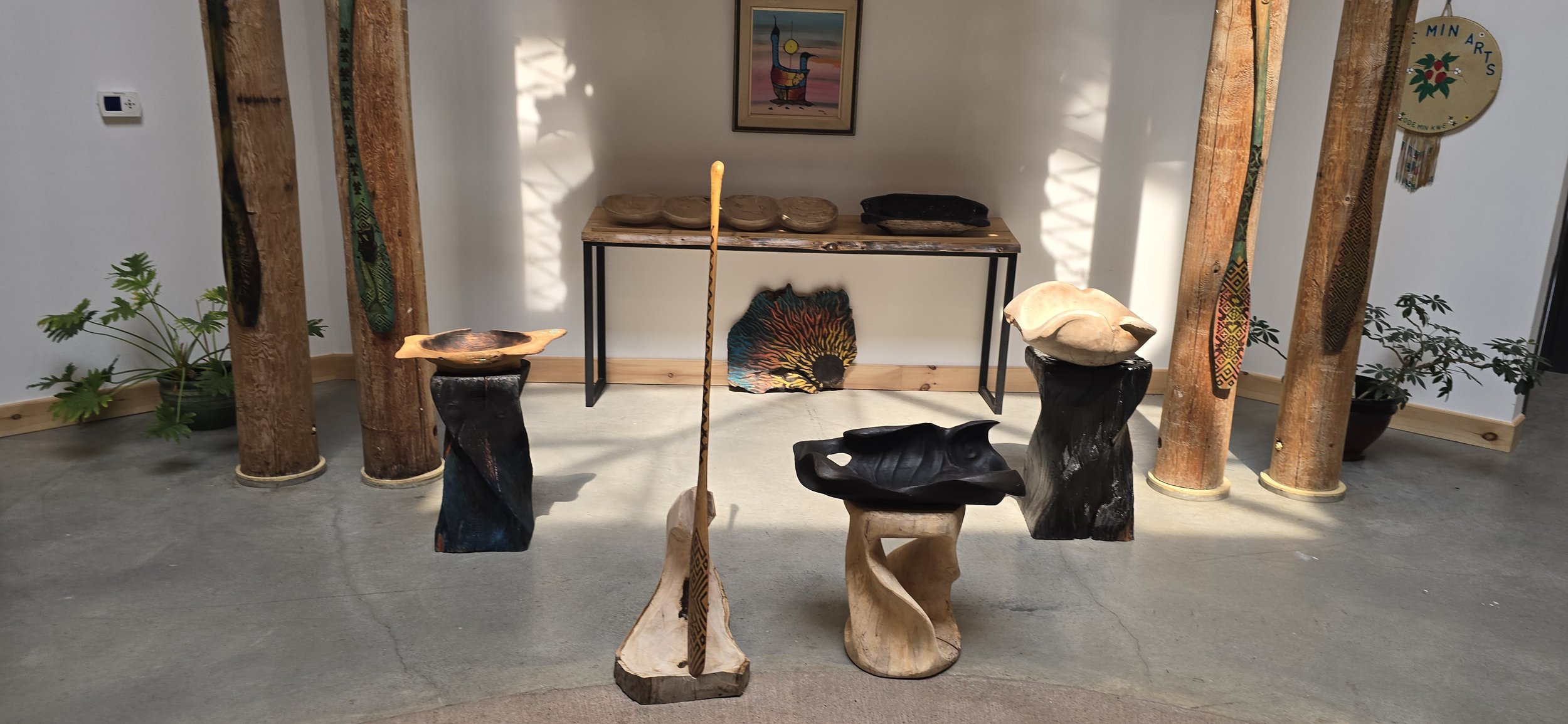 Wooden sculptures and bowls displayed in a gallery with a framed painting on the wall, plants on each side, and wooden columns as decor.