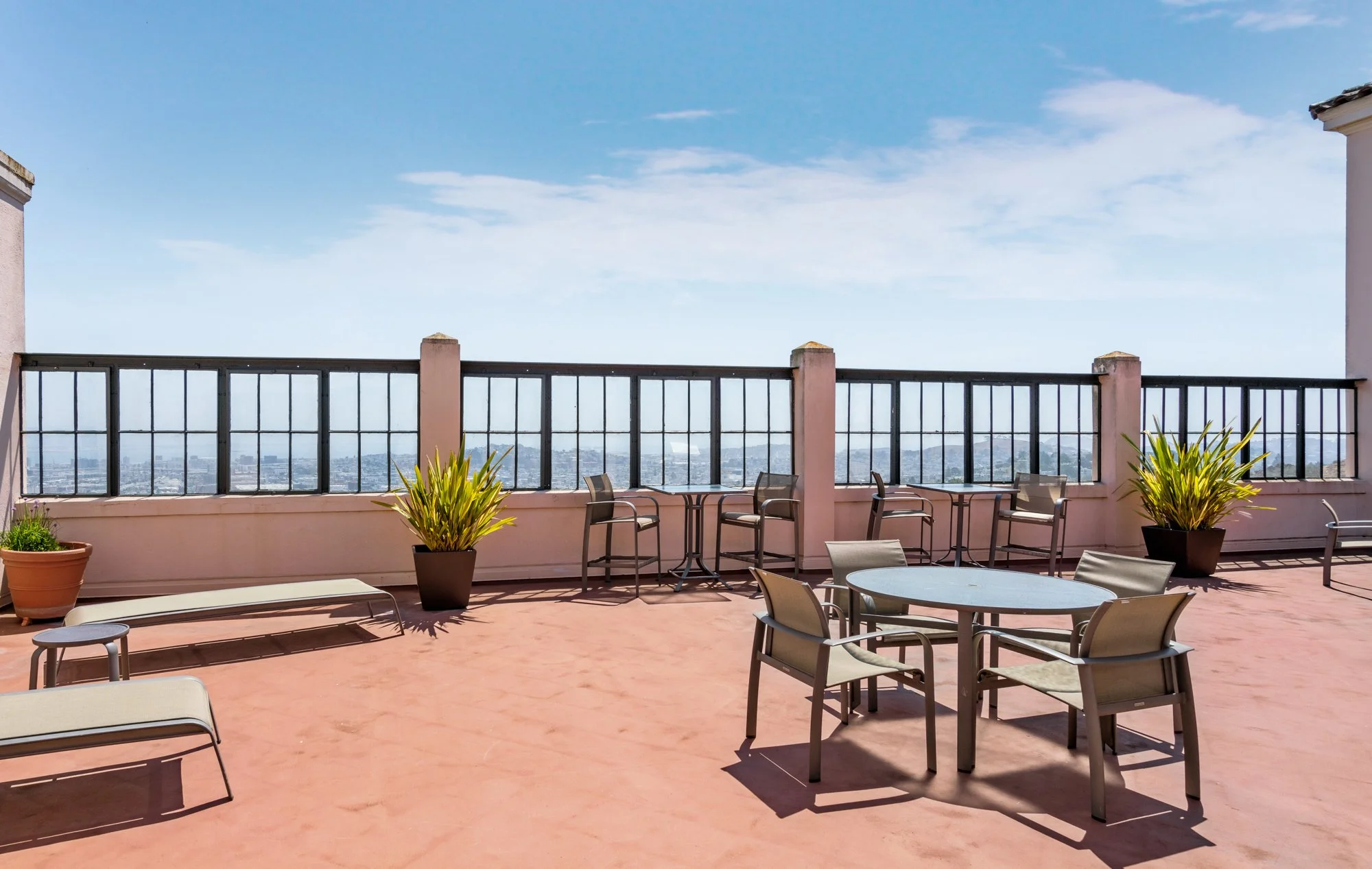 Rooftop terrace with a pink floor, black railing, potted plants, glass tables, and chairs against a city skyline and blue sky with clouds.