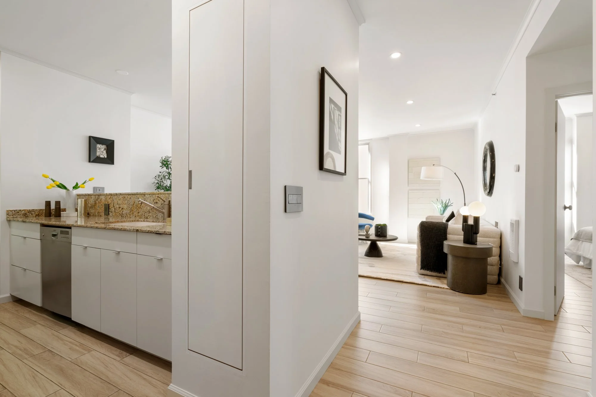 Open-concept living room and kitchen with white walls, hardwood floors, modern furnishings, and decorative lighting.