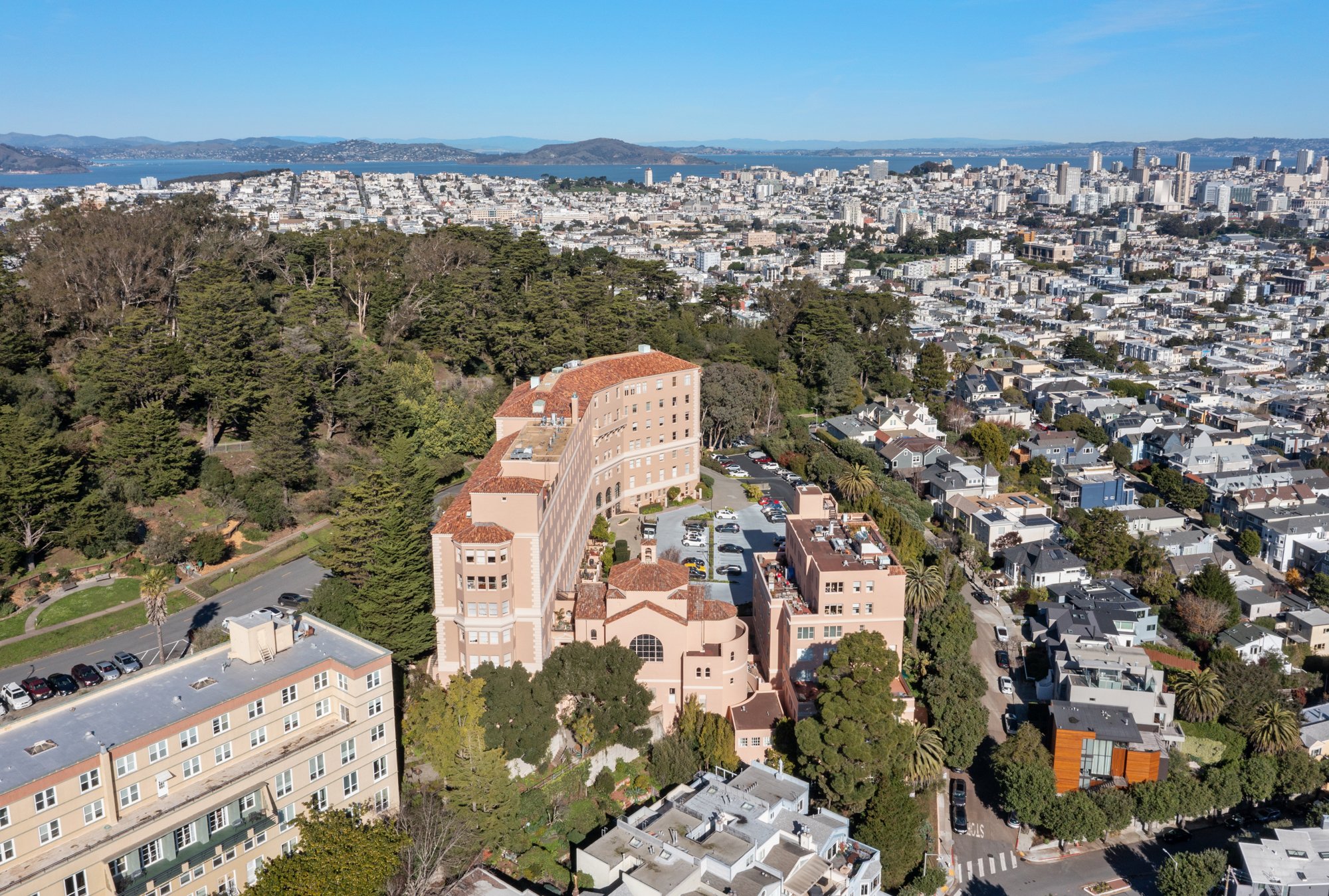 Aerial view of San Francisco with a hotel, residential houses, green trees, and cityscape with water and distant hills in the background.