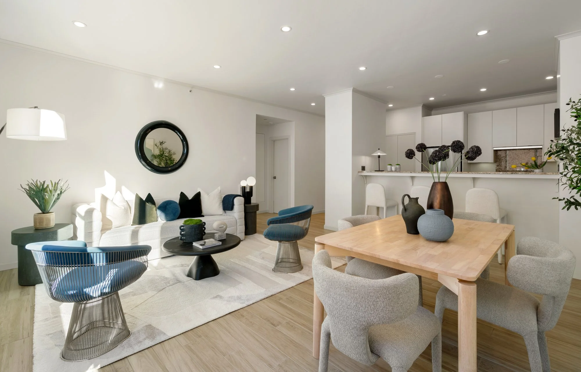Modern living room and dining area with white walls, light hardwood floors, a white sofa with pillows, a black round coffee table, blue and black chairs, a wooden dining table with four beige chairs, and decorative vases on the table. The kitchen is 