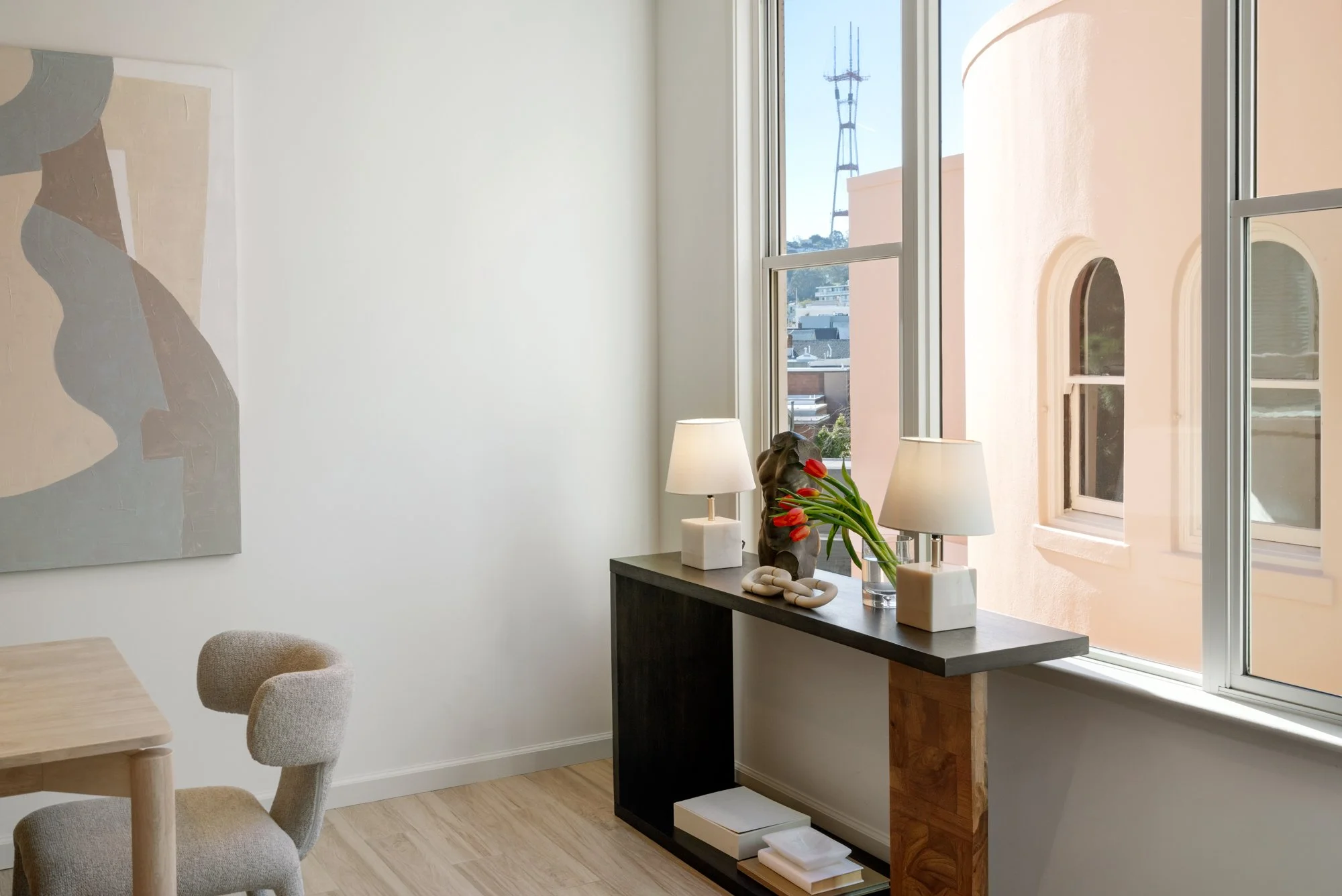 A bright interior with a table, a chair and a black console table near a large window. The console table holds two white lamps, a vase with red tulips, a decorative sculpture, and books underneath. The wall has a large abstract painting, and outside the window, a building and a tall transmission tower are visible.