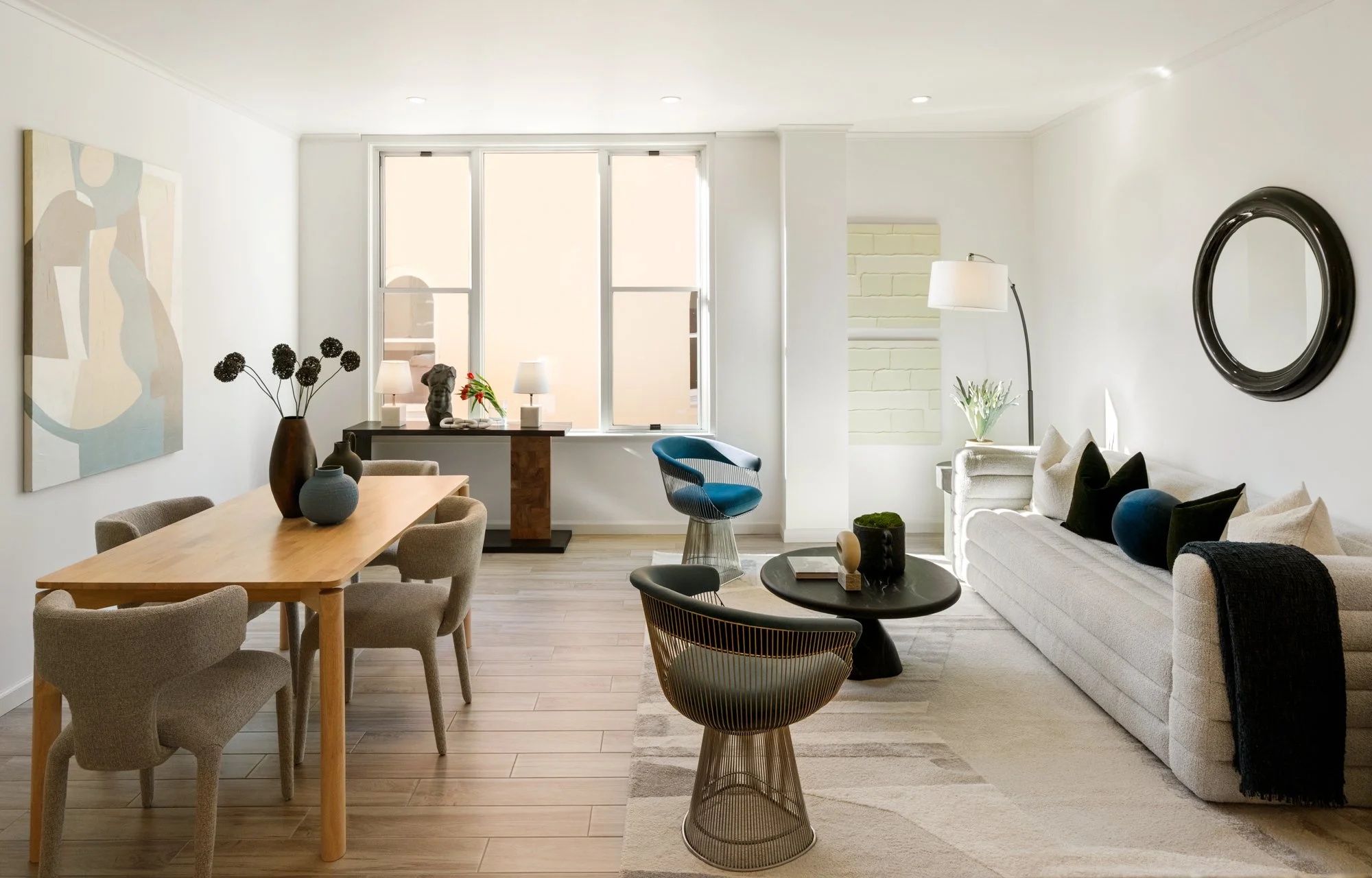Modern living room with wooden dining table, beige chairs, white sofa, and decorative black pillows. Artwork on wall, and various vases with plants and flowers.