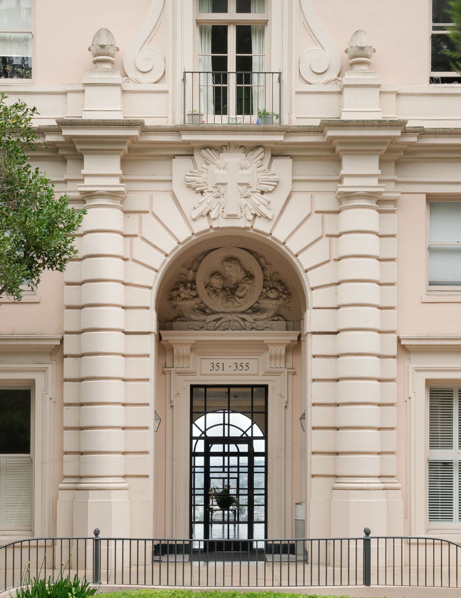 Facade of a beige-colored building with ornate architectural details, archway entrance, decorative carvings, and a glass door leading inside.