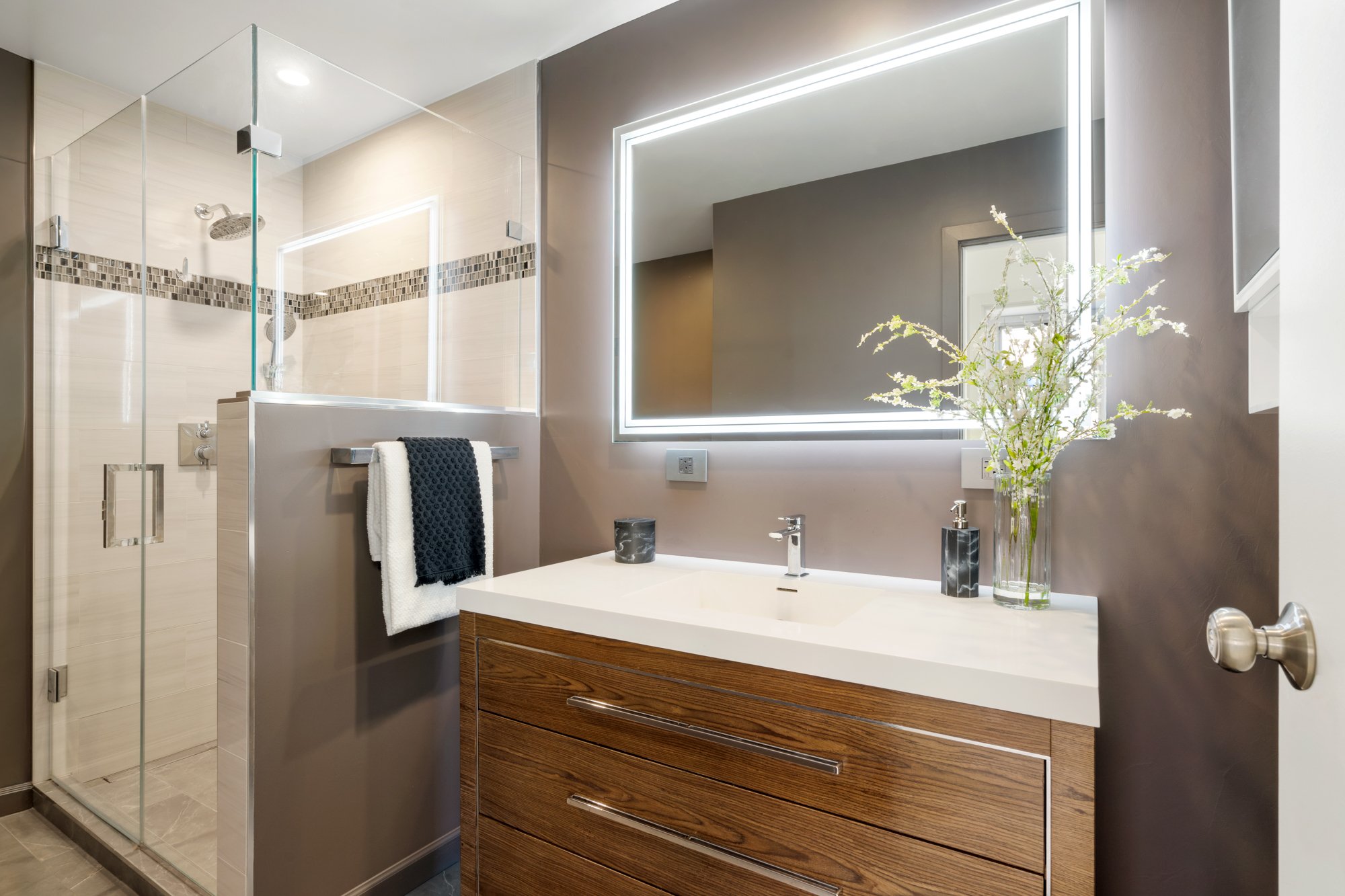 Modern bathroom with glass shower enclosure, illuminated rectangular mirror, wooden vanity with drawers, vase with flowers, and black soap dispenser.