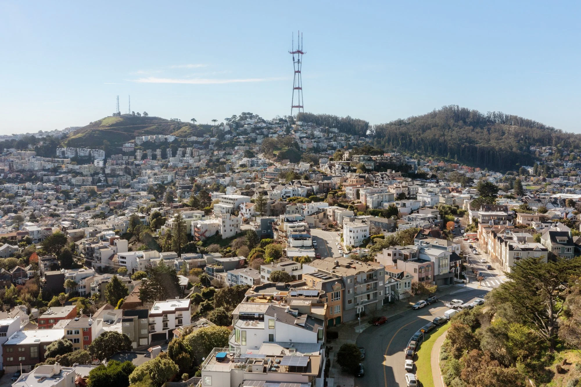 Aerial view of a hilly urban neighborhood with a large radio tower on top of the hill, surrounded by houses, trees, and a clear blue sky.