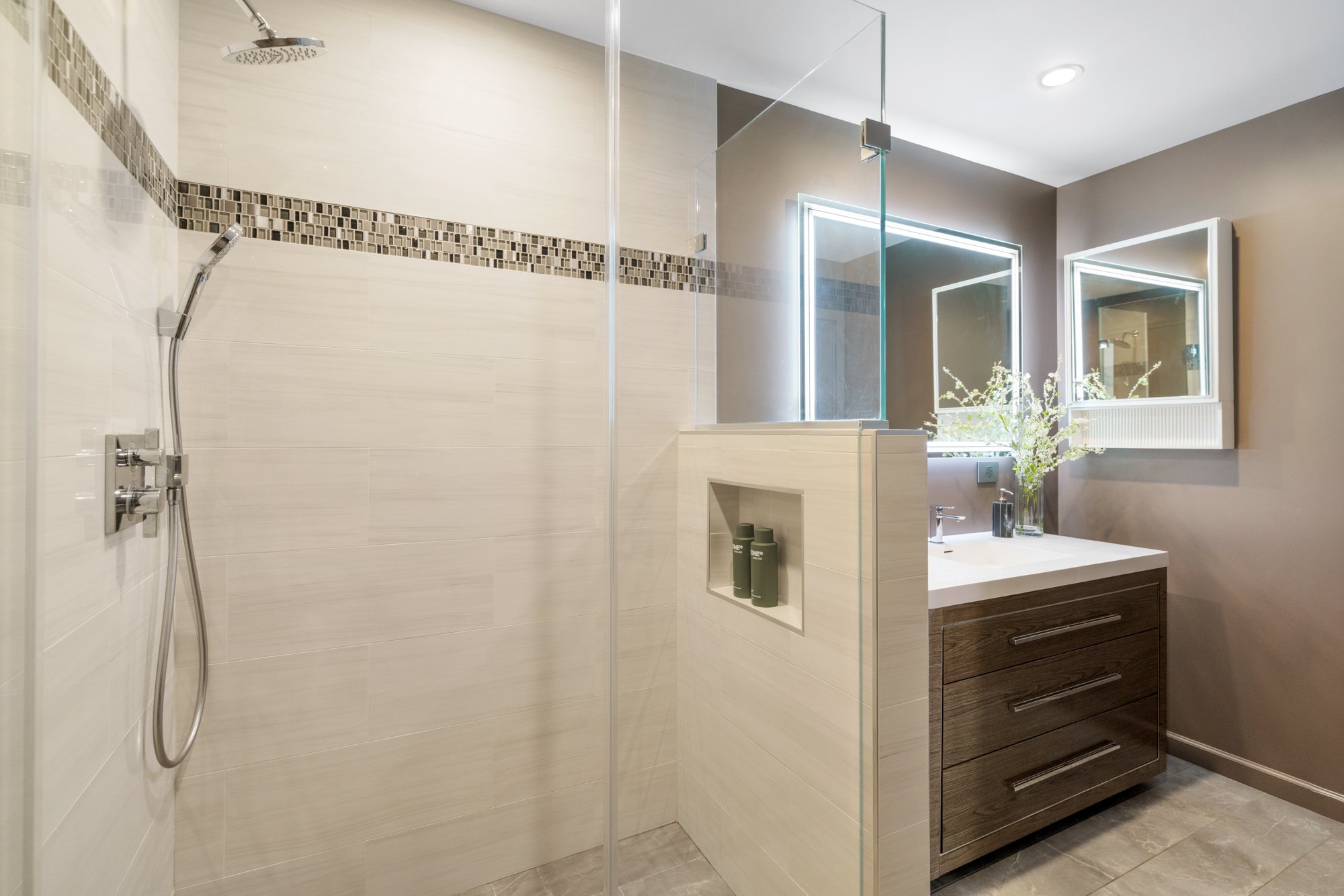 Modern bathroom with a walk-in shower featuring a glass door, wall-mounted showerhead, and handheld shower. A dark wood vanity with three drawers and a white countertop, a double mirror with LED lighting, gray walls, and beige tiled flooring.