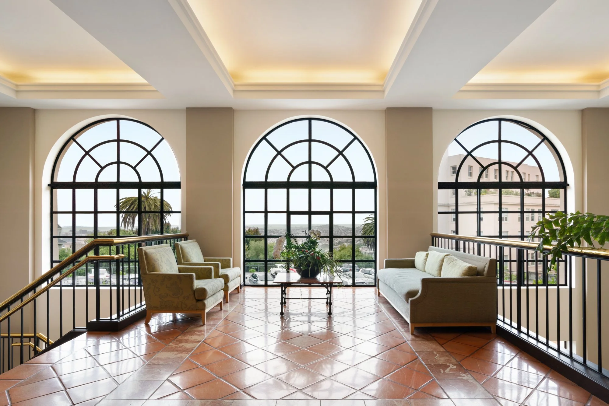 Interior of a bright lounge area with large arched windows, furniture including two armchairs and a sofa, a small coffee table with a plant, and city view outside