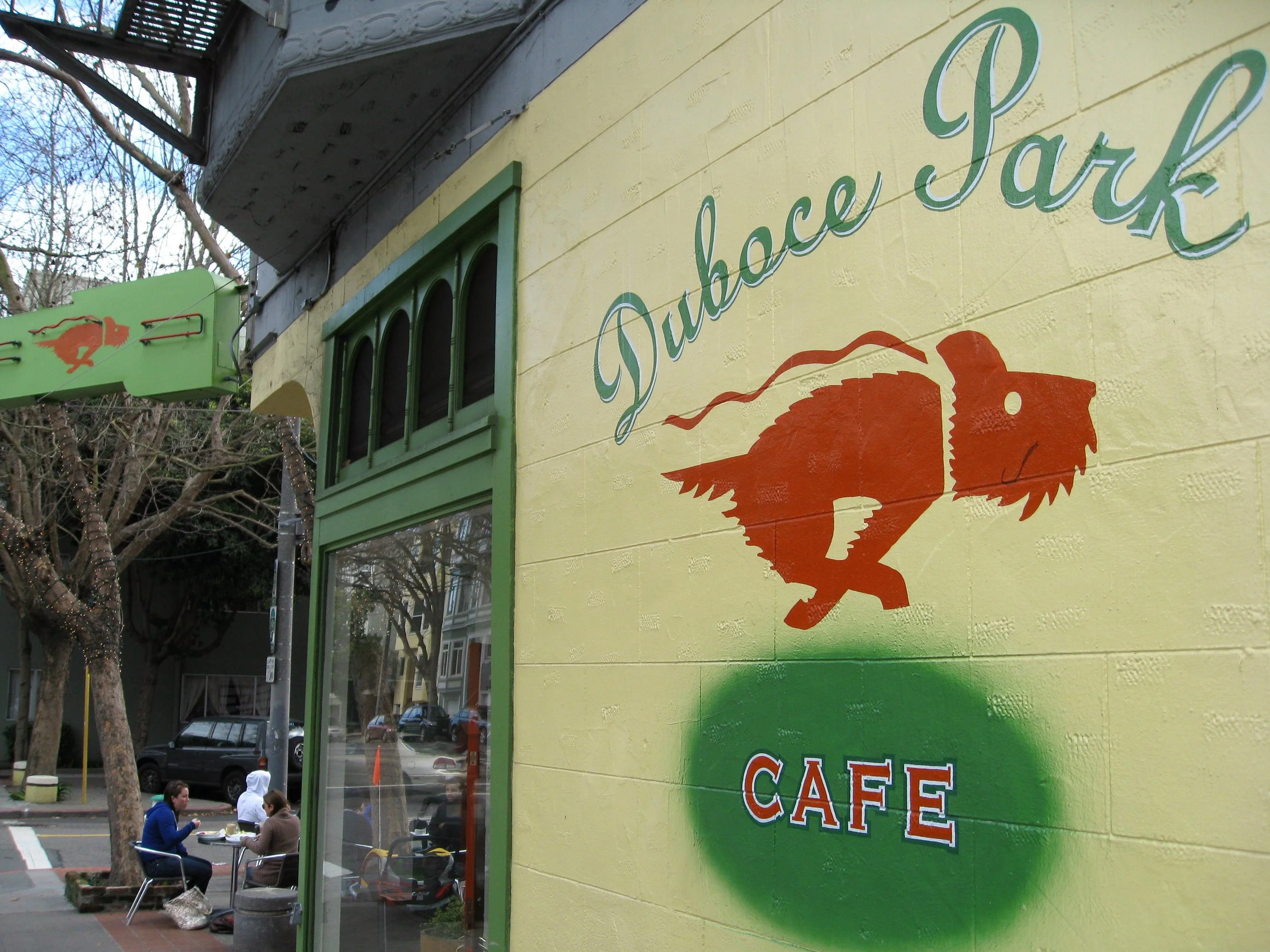 Exterior wall of Duboce Park Café with a painted logo of a dinosaur with a chainsaw in orange, and the words 'Duboce Park' and 'Café' painted on the yellow wall.