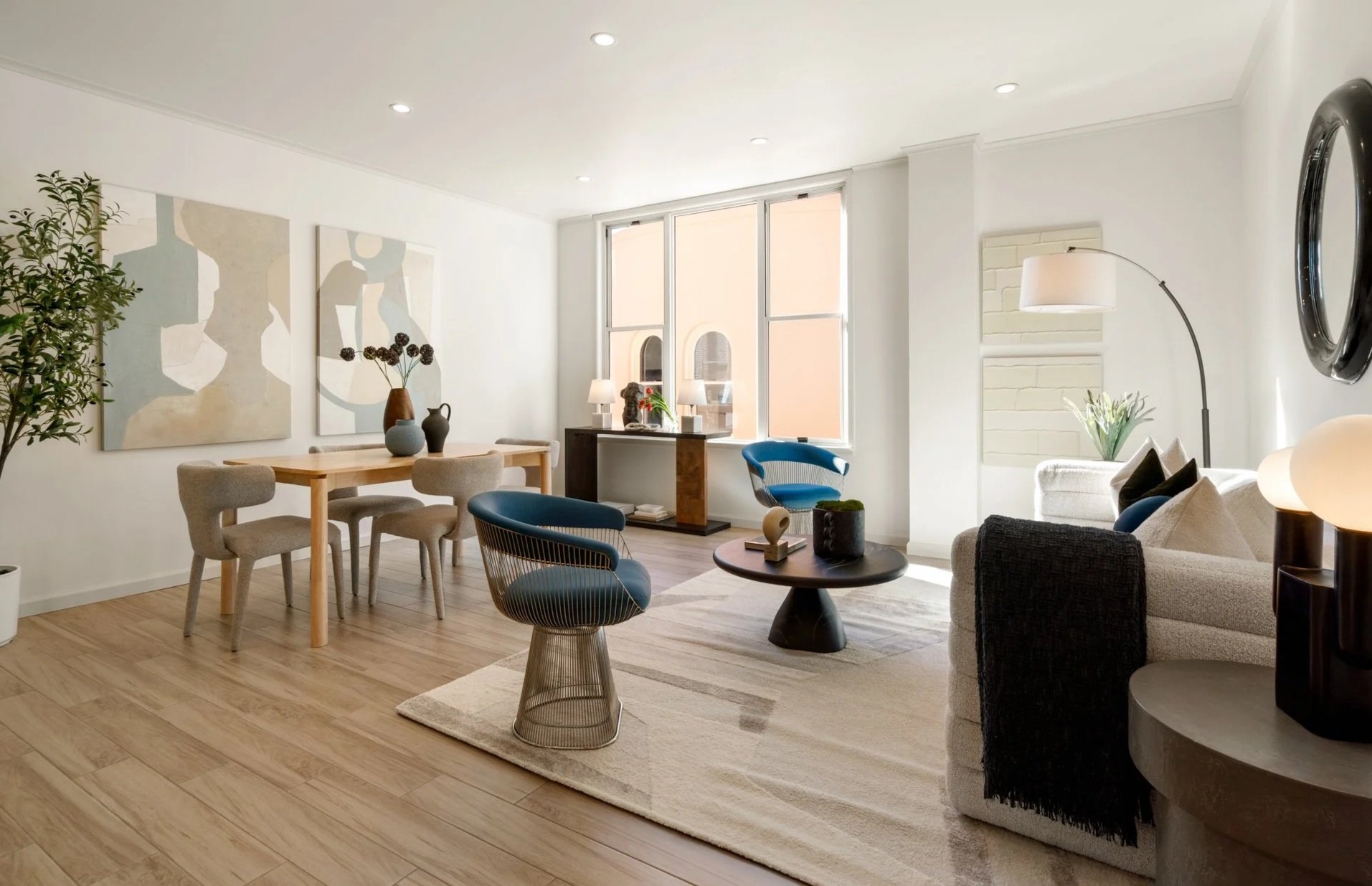 Bright living room with white walls, wooden flooring, a white sofa with black and beige pillows, and modern blue and black chairs around a dark coffee table. Artwork decorates the walls, and large windows let in natural light.
