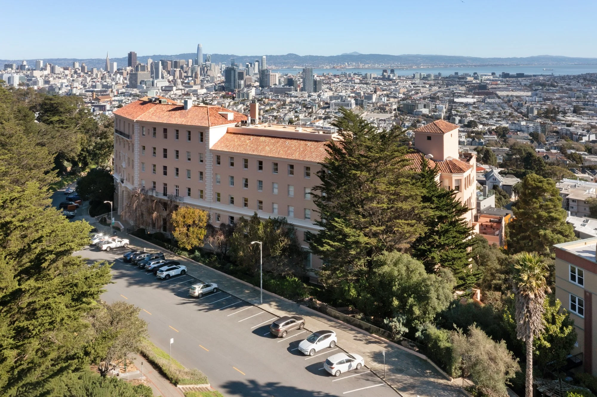 City skyline with tall buildings and a bay in the background, viewed from a hilly area with trees and a parking lot in the foreground.
