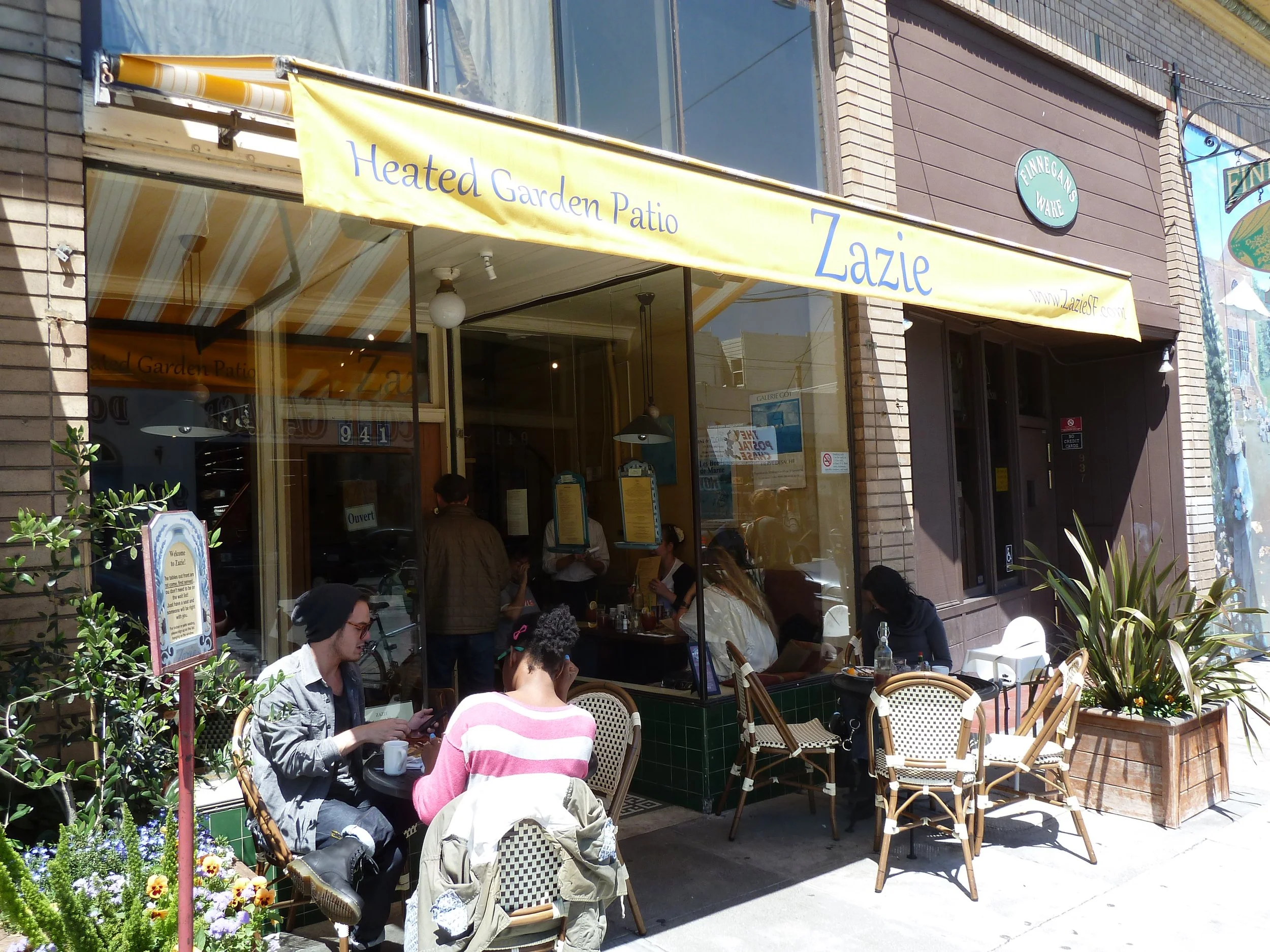 Restaurant named Zazie with outdoor seating under a yellow awning reading 'Heated Garden Patio.' Several customers are sitting outside, and more are visible inside through large windows. There are potted plants and flowers outside.