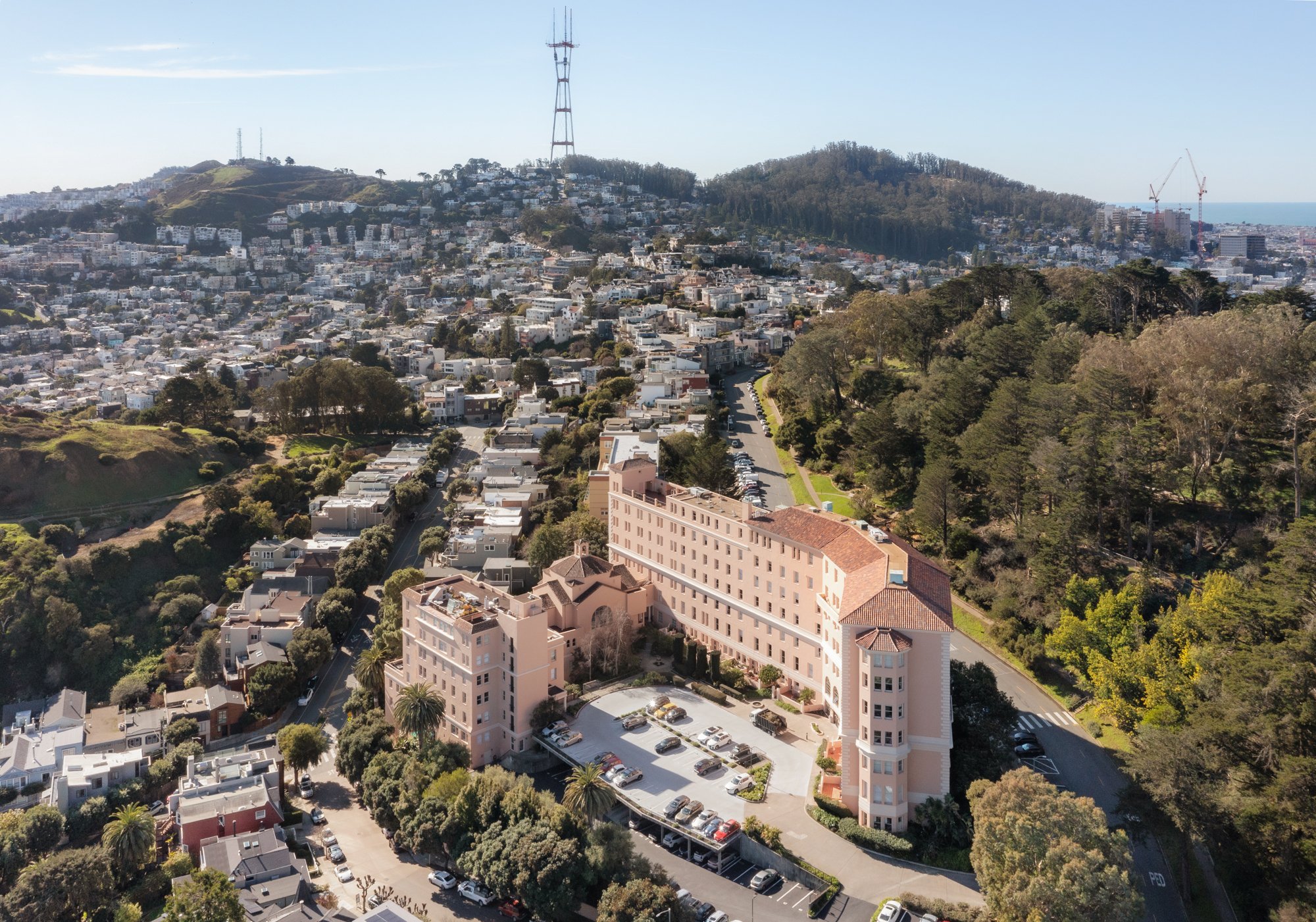 Aerial view of a large, historic pink building with a parking lot in front, surrounded by lush green trees and residential houses, with hills and cityscape in the background.