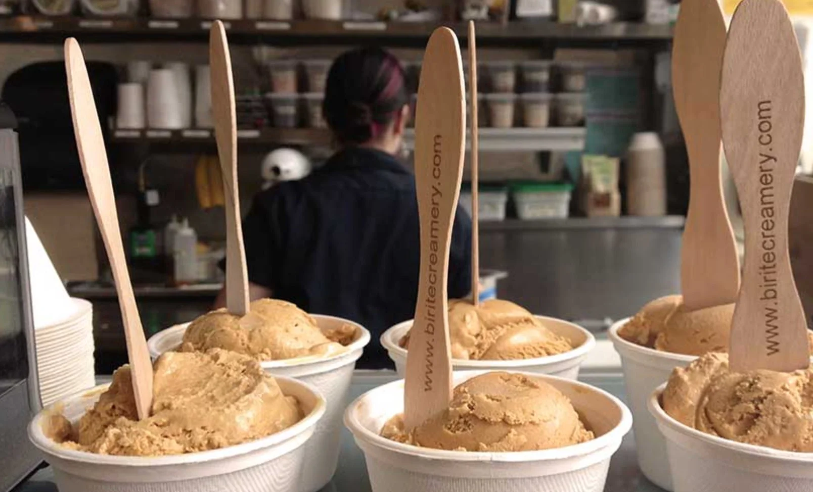 Six cups of caramel ice cream with wooden spoons labeled www.bitecreamery.com on a counter in an ice cream shop, with a person working in the background.
