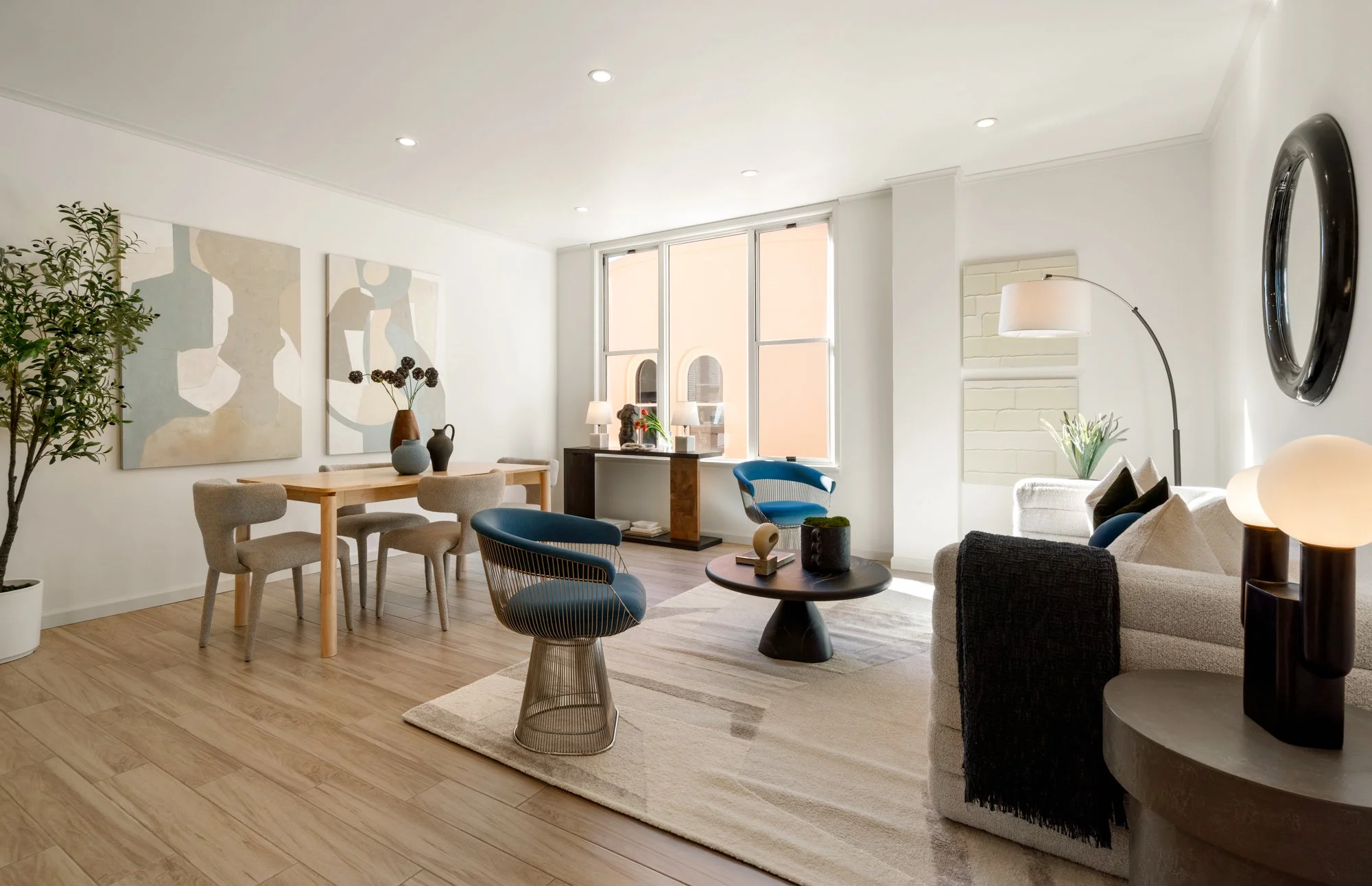 Bright living room with white walls, wooden floors, a white sofa with dark cushions, a coffee table, and modern chairs, featuring art on the walls and large windows allowing natural light.