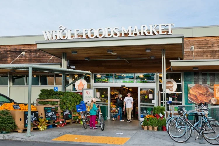 Exterior of Whole Foods Market with shoppers entering, bicycles parked outside, and fresh produce displayed near the entrance.