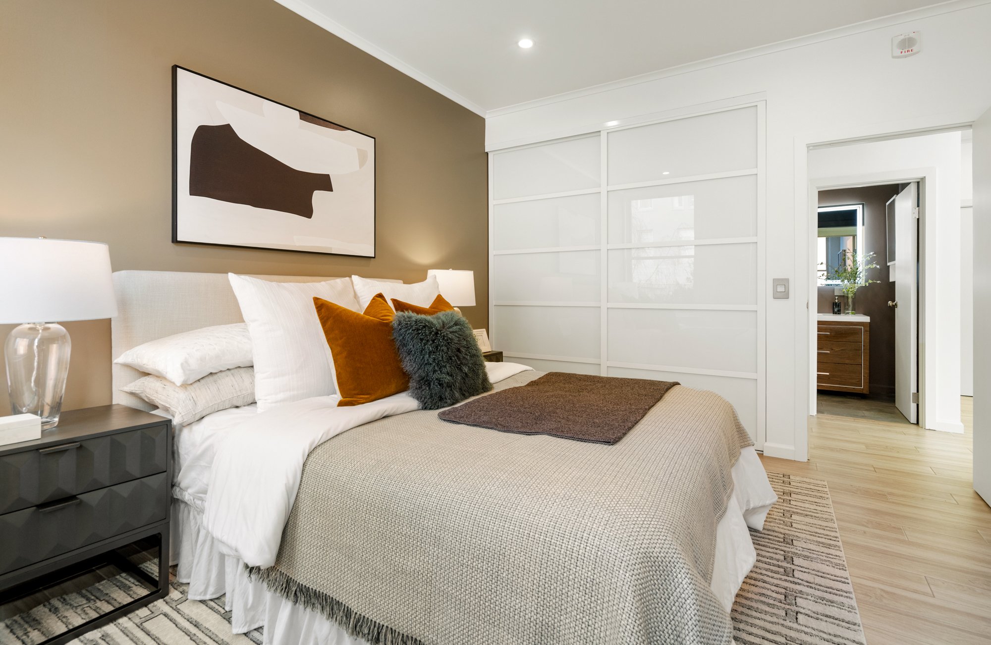 Modern bedroom with beige and white decor, abstract wall art, white bed with brown and black pillows, nightstand with lamp, sliding door closet, and adjacent bathroom with wooden vanity.