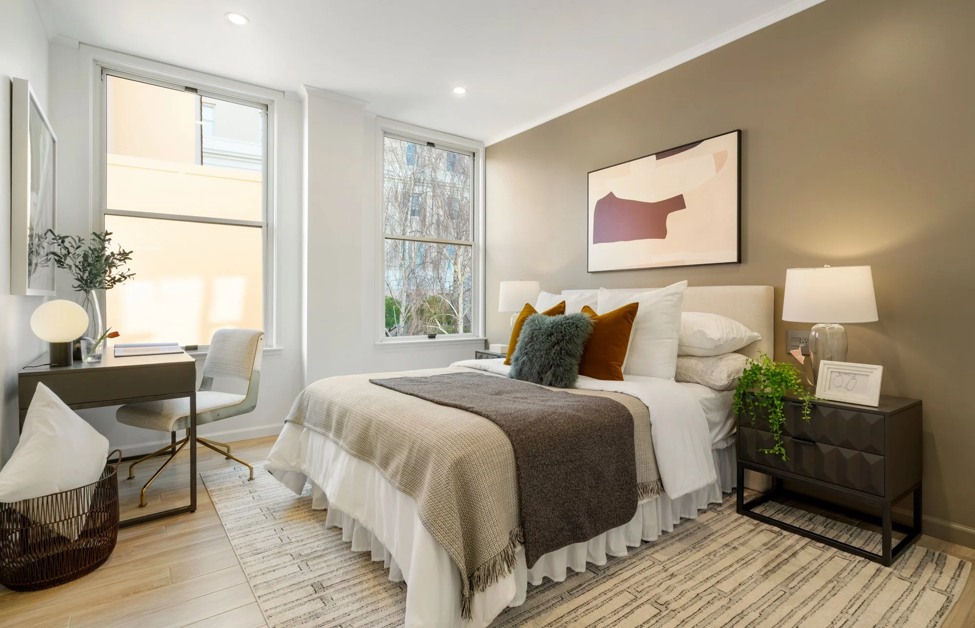 Modern bedroom with a bed covered in white linens, a gray and brown throw blanket, and multiple decorative pillows, flanked by matching bedside lamps, with large windows showing leafless trees outside, and a small workspace with a desk and chair near
