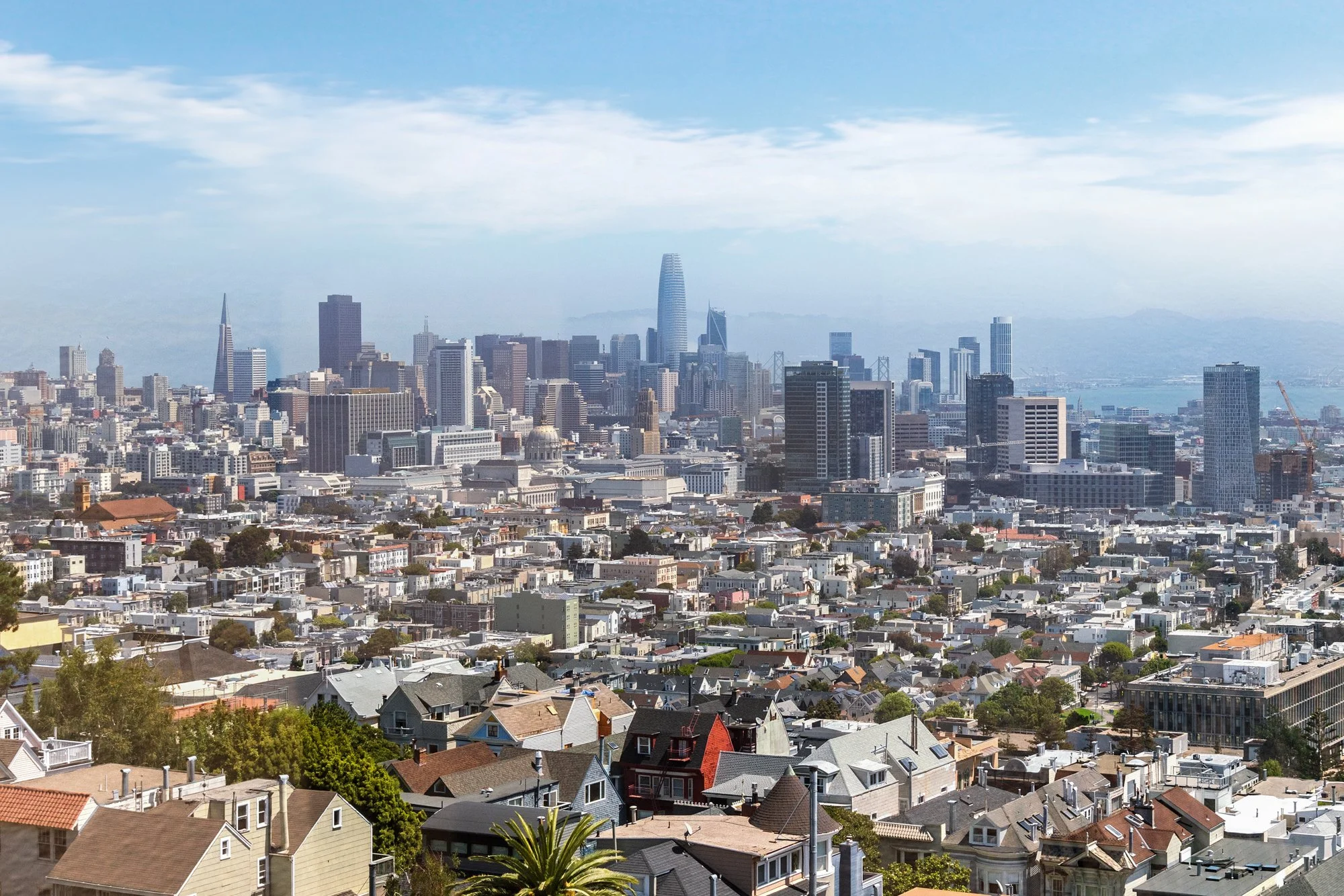 City skyline of San Francisco with skyscrapers, residential houses in foreground, and blue sky with some clouds.