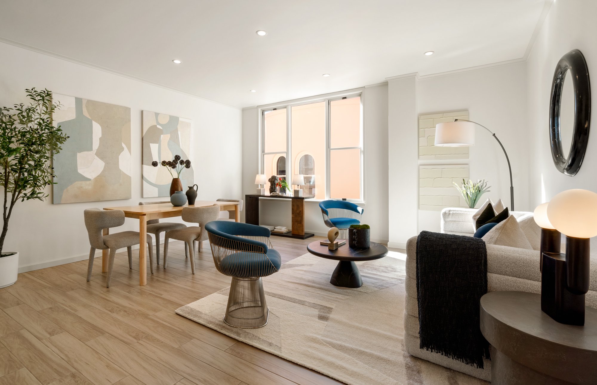 Bright living room with white walls, wooden flooring, and modern furniture. Features a white sofa with dark cushions, a round black coffee table, and blue chairs. Large windows with sunlight, artwork on the walls, and various decorative elements like