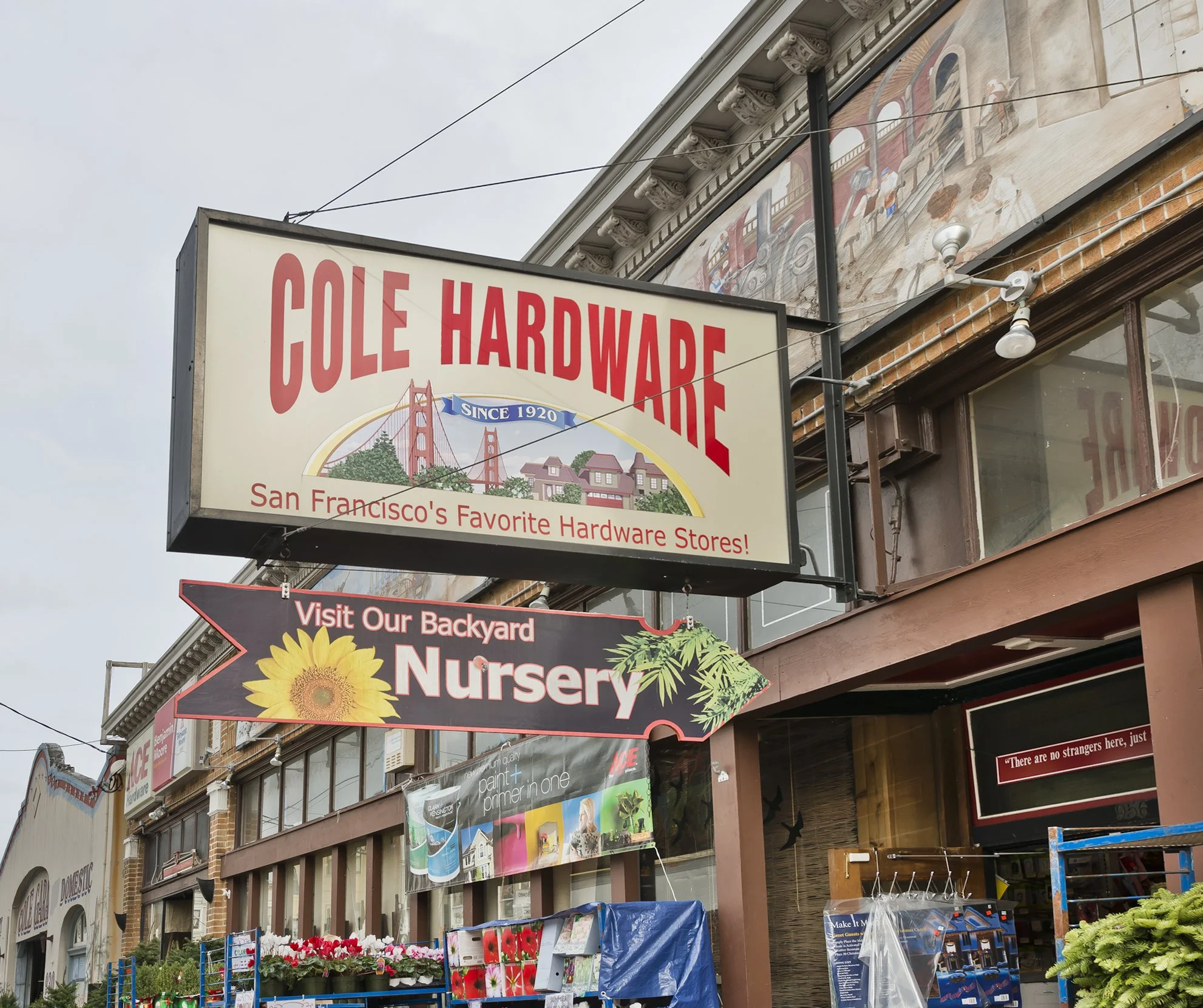 Photo of a storefront with signs for Cole Hardware and a backyard nursery, with flower plants displayed outside.