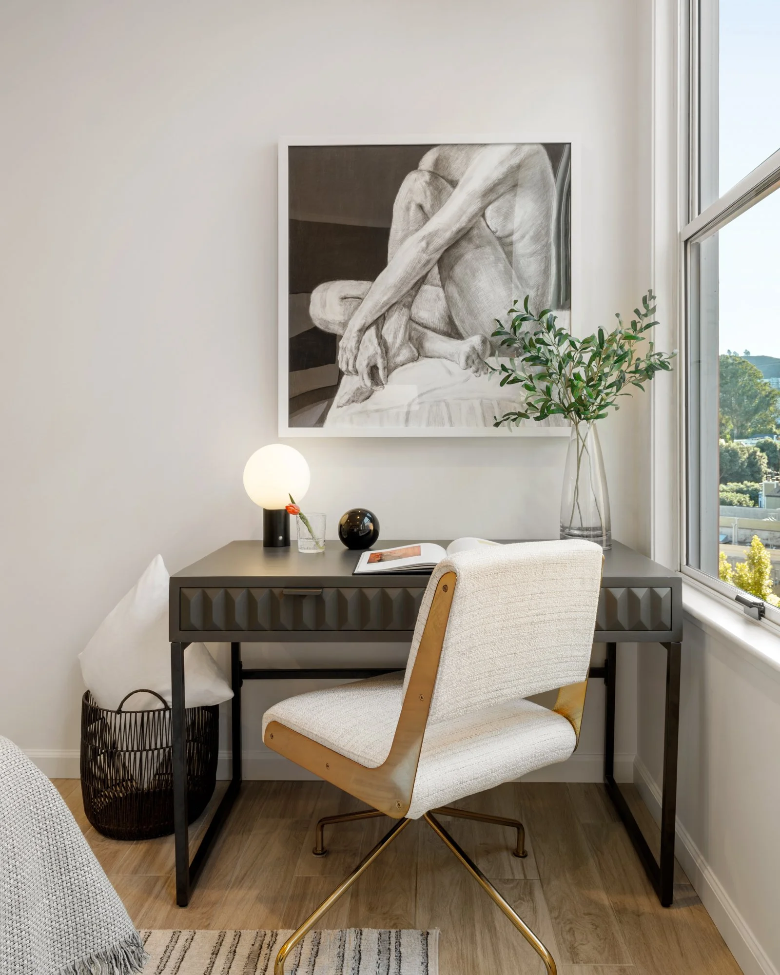 A modern home office with a black desk, white upholstered chair, and a black and white artwork of a nude figure on the wall. A green plant in a glass vase is on the desk, and a round white lamp and black decorative ball are also present.