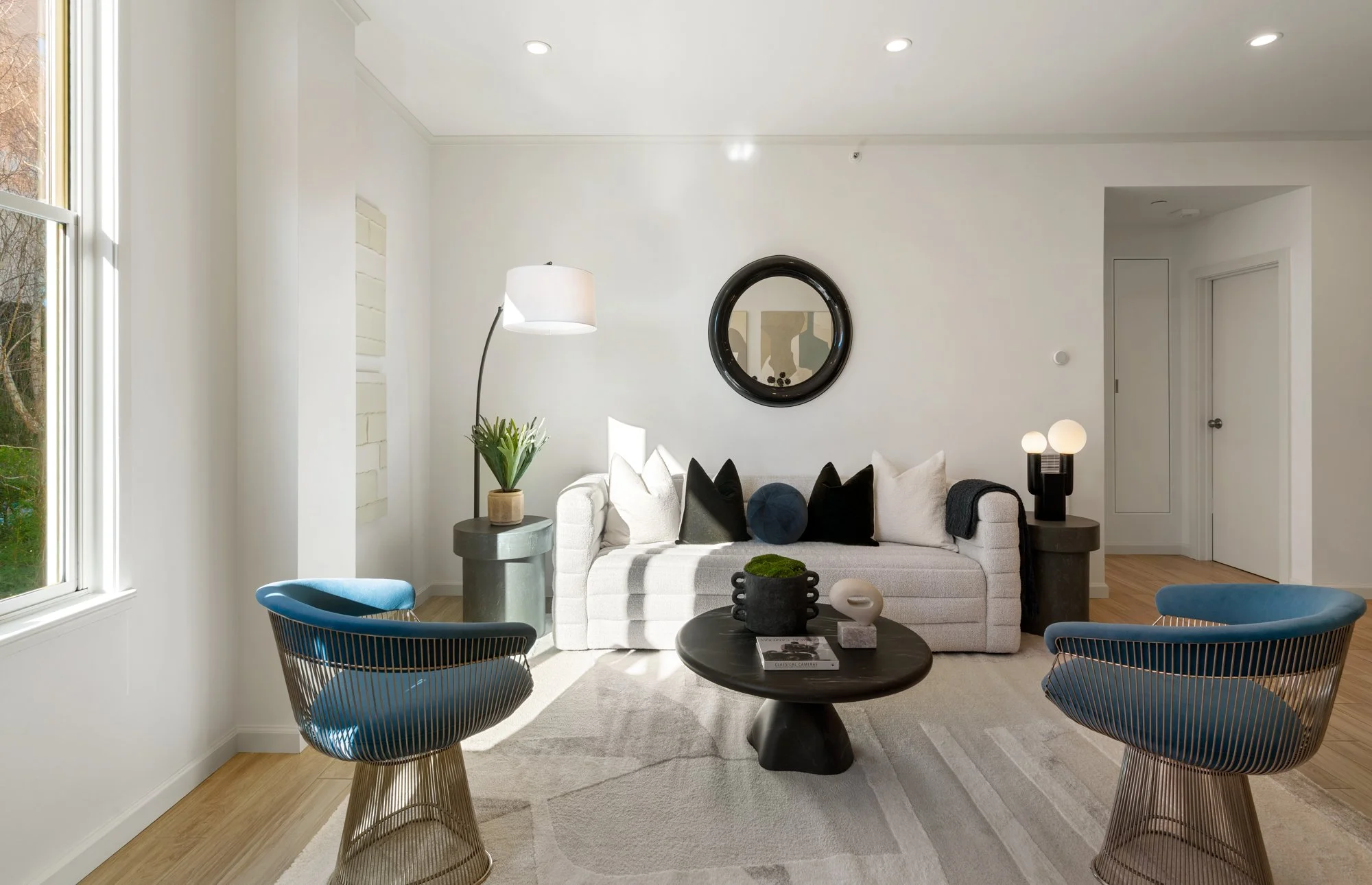 Modern living room with white sofa, blue chairs, coffee table, and large window with sunlight.