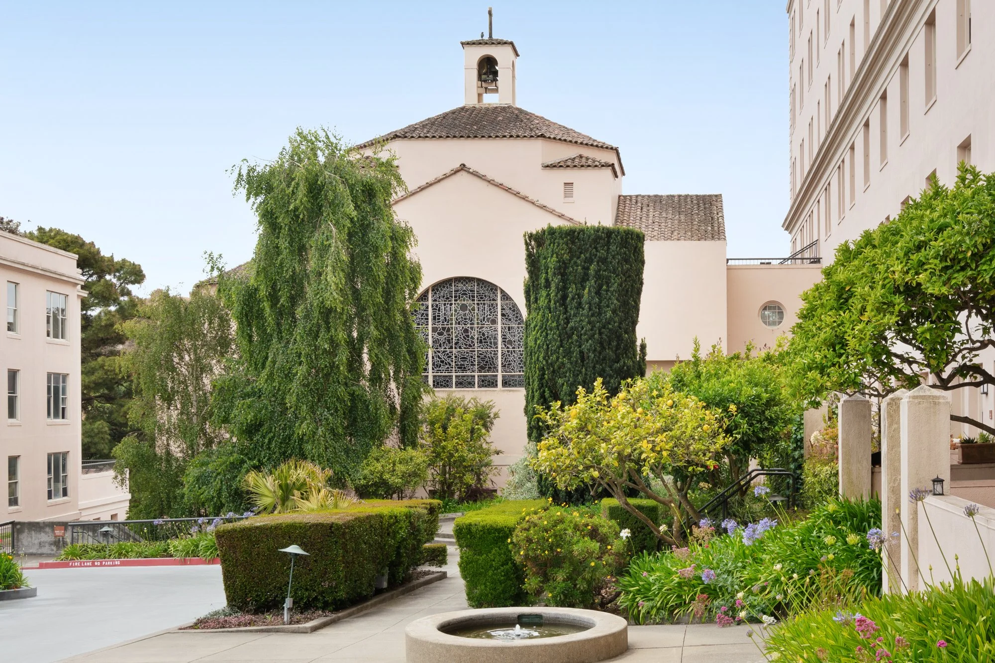 A small church with a bell tower and stained glass window, surrounded by lush greenery and flowering plants, with adjacent buildings and a paved courtyard in the foreground.