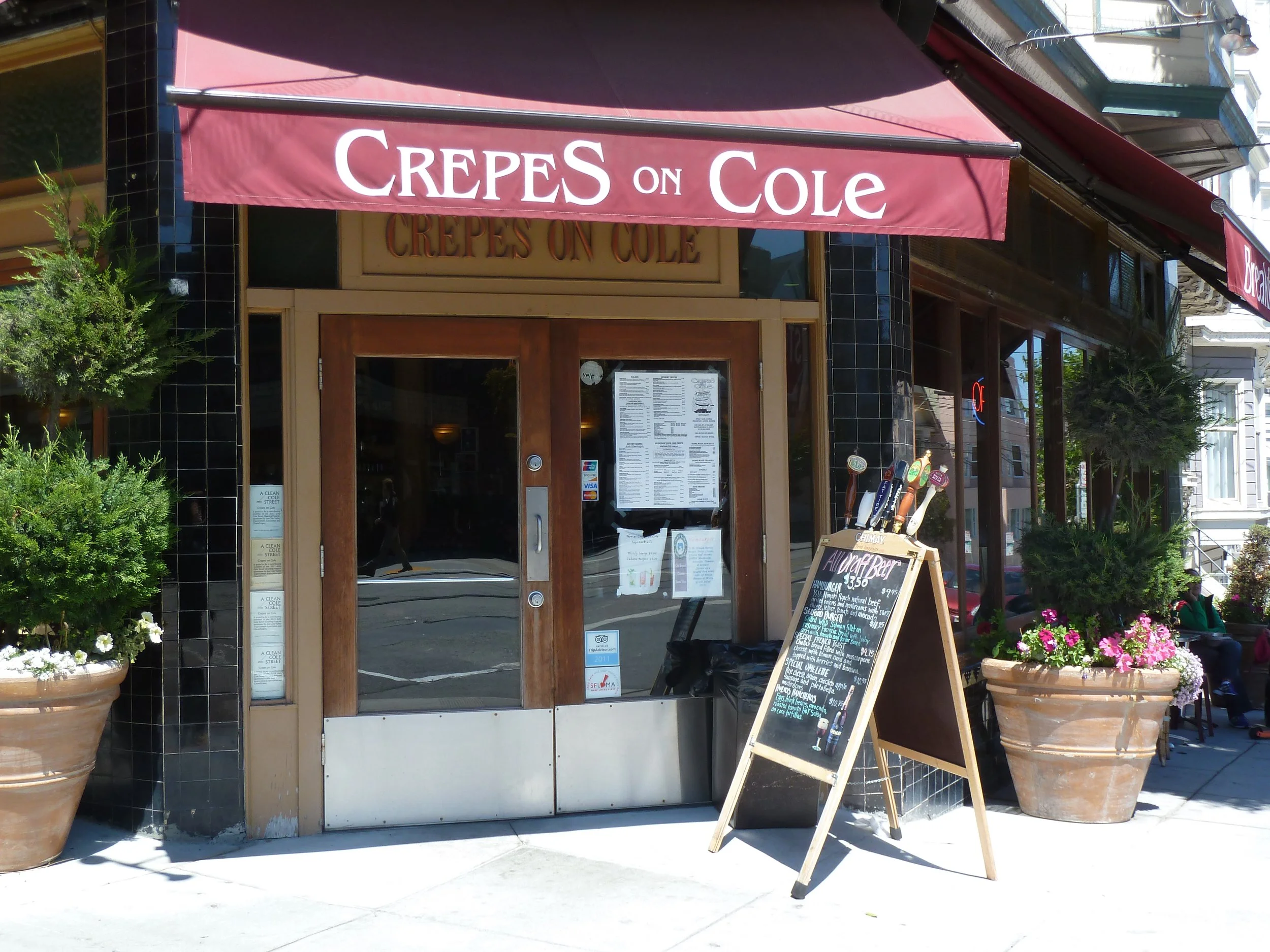 Exterior of a restaurant called 'Crepes on Cole' with a maroon awning, potted plants on the sidewalk, and a chalkboard menu outside.