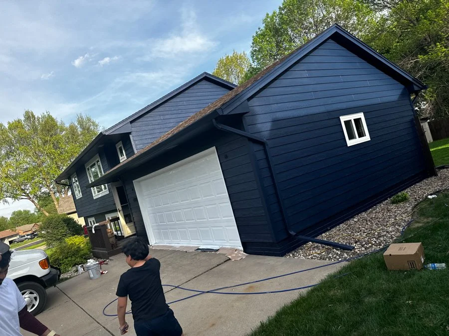 A newly painted dark blue house with white garage door, two windows, and a sloped roof, with trees and a partly cloudy sky in the background. Two boys are on the driveway, one holding a hose, with a cardboard box and outdoor supplies nearby.