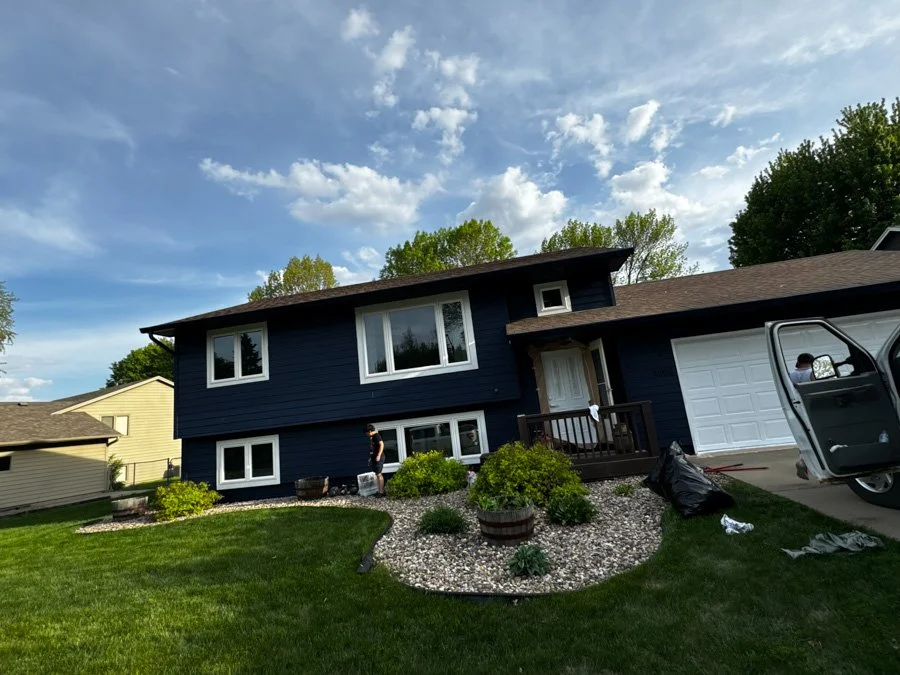 A two-story dark blue house with white window frames, a small front porch, and a white garage door. A person is standing near the house, and another is partially visible in a van. The front yard has green grass, a landscaped flower bed with bushes an