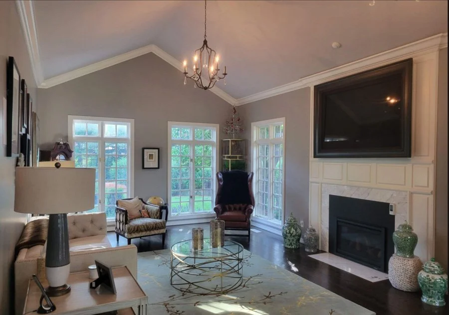 Living room with gray walls, large windows, chandelier, and fireplace with mounted TV. Includes various chairs, a glass coffee table, and decorative vases.
