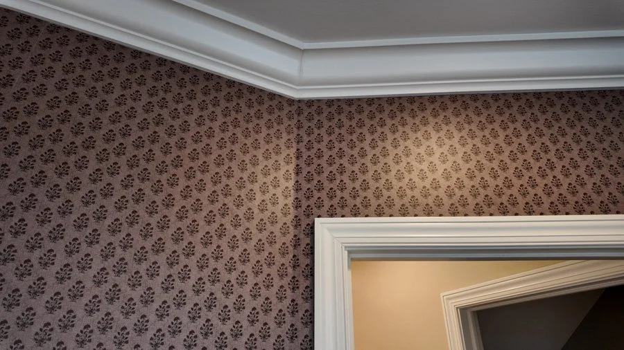 Corner of a room showing patterned wallpaper, crown molding, and a door frame.