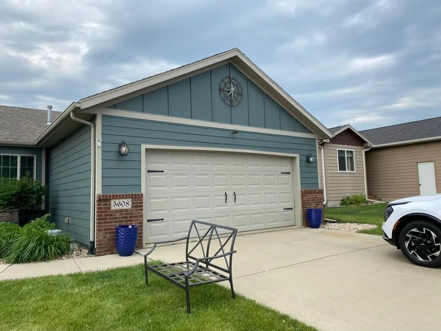 Front view of a suburban house with a two-car garage, modern light fixture, blue planters, a bench, and a white car parked in driveway under a cloudy sky.