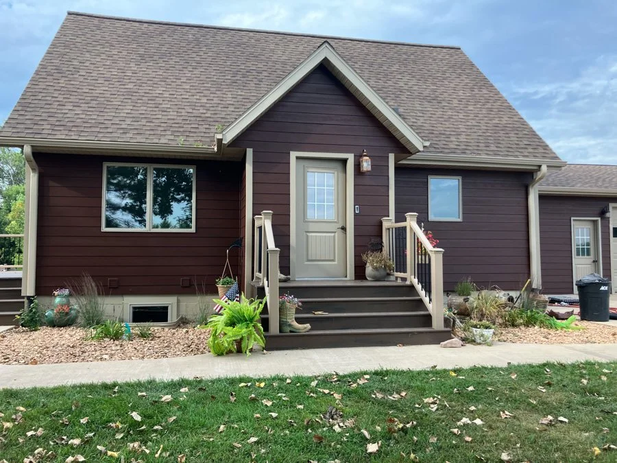 A small house with dark brown siding, a gray door, and a gable roof, surrounded by a well-kept yard with a sidewalk, plants, and decorative items.