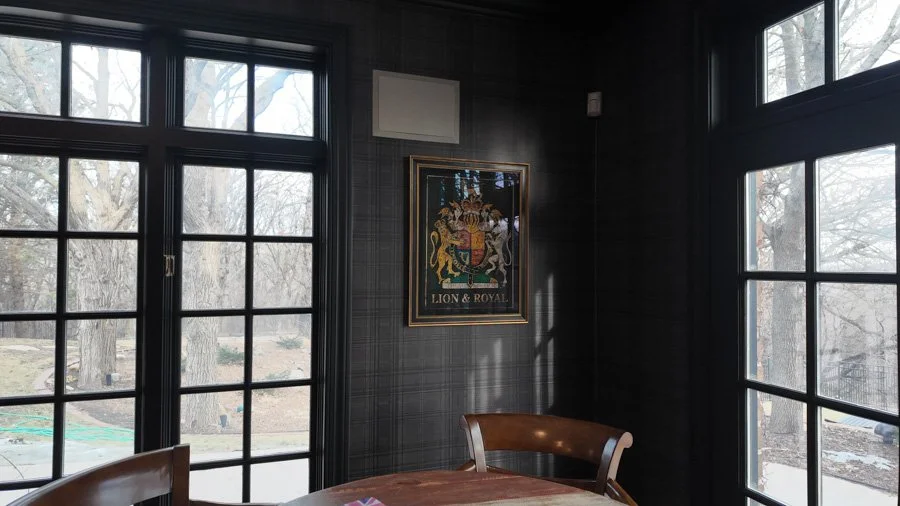 Interior of a room with dark plaid walls, large windows, a framed coat of arms crest with the words 'Lion & Royal,' a wooden table, and a chair.