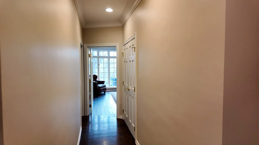 A hallway leading to a living room with large windows, a chair, and hardwood flooring.