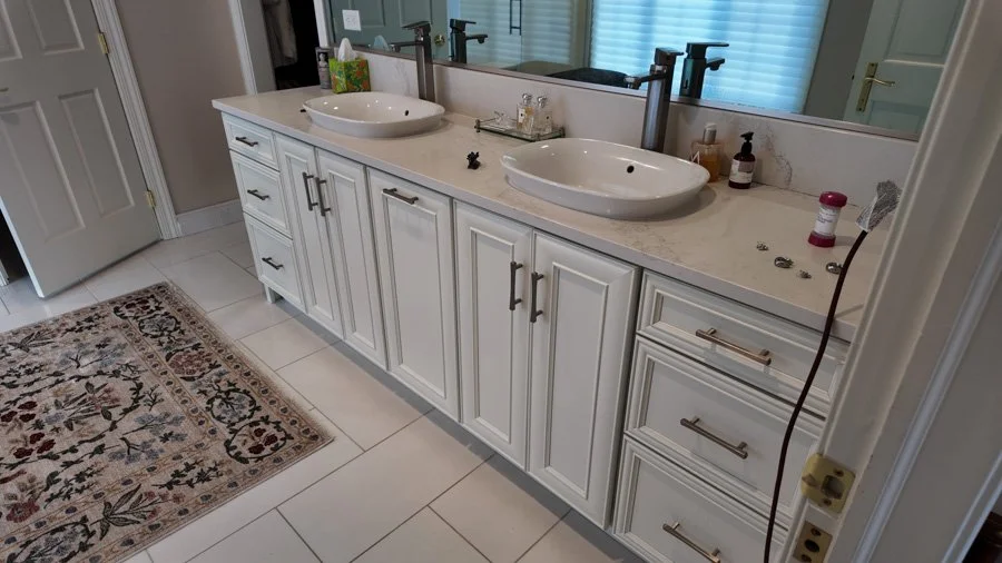Bathroom vanity with double vessel sinks, white cabinetry, a large mirror, and various toiletries on the countertop.