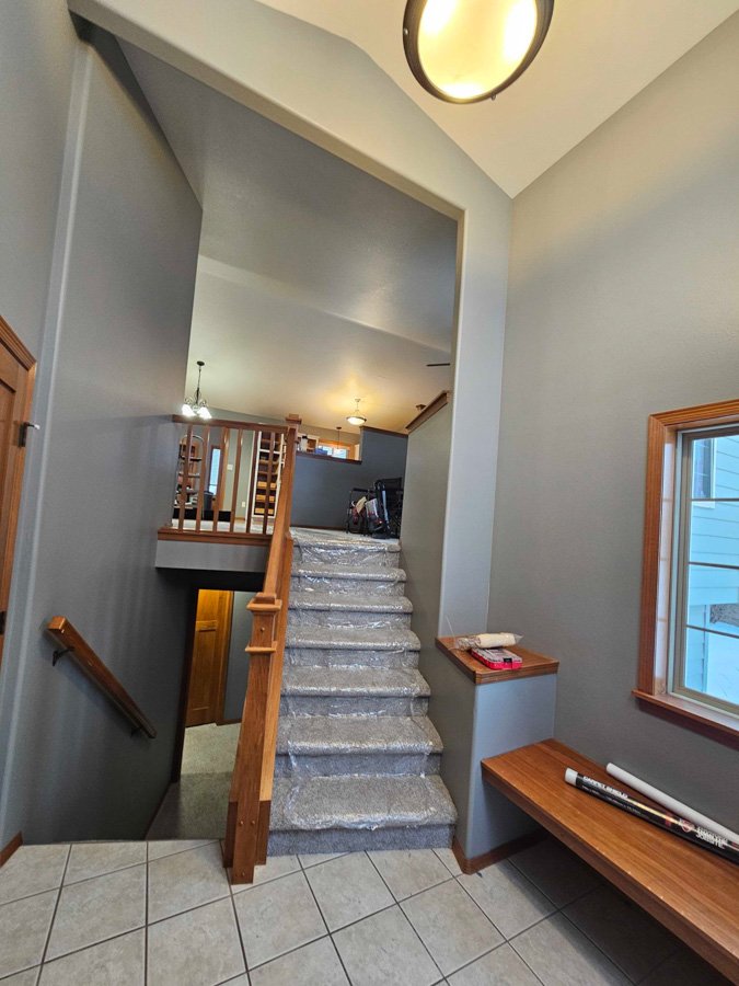 Interior view of a house showing a staircase with carpeted steps leading to the second floor, wooden handrails, a tiled floor, a small wooden bench, and a window with wooden trim.