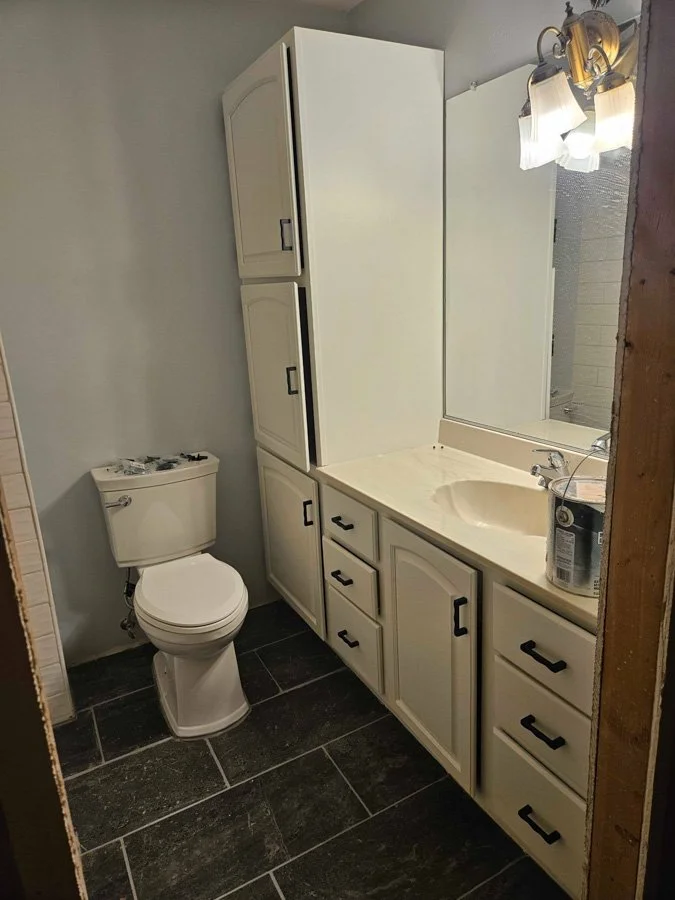 Small bathroom with white toilet and vanity with sink, black hardware, large mirror, light fixture, black tiled floor, and a bucket on the counter.