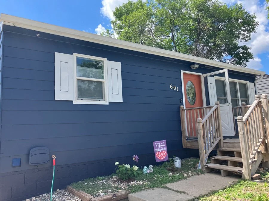 A blue house with white window shutters, a wooden front porch with stairs, and a red front door with an oval glass insert. There are a few small plants and decorations in the front yard, with a sign that reads 'A Home for Heart.'