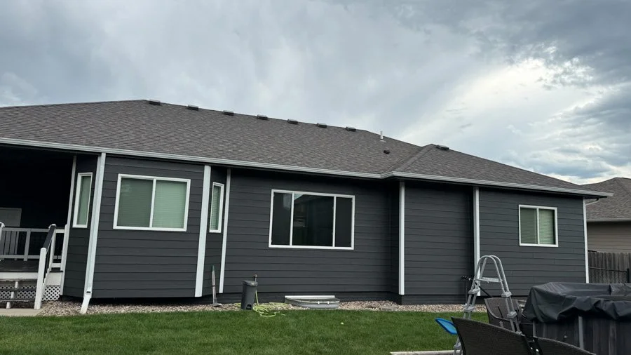 Rear view of a modern gray house with a sloped roof and several windows, beyond a green lawn with patio furniture and an overcast sky.