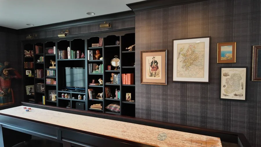 Living room with black bookshelf filled with books and decor, framed artwork on plaid wallpaper, and a track light ceiling fixture.