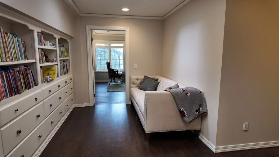 A living room with a white bookshelf filled with books on the left, a beige sofa with pillows and a blanket on the right, and a doorway leading to a home office with a window and a black office chair.