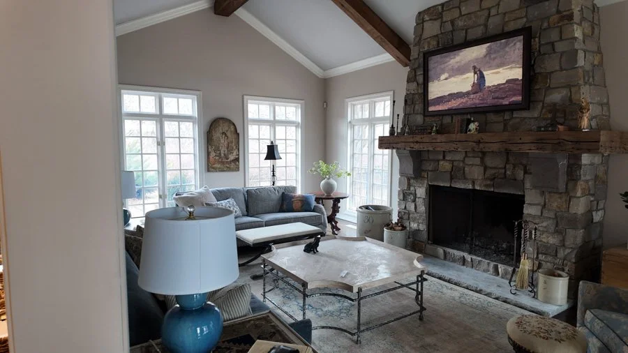 Living room with a stone fireplace, a sofa, coffee table, side tables, lamps, and large windows with white framing, decorated in neutral and earthy tones.