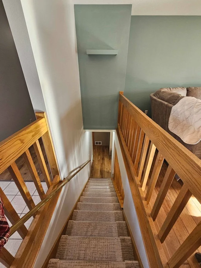 View looking down a staircase with light-colored carpet, wooden handrails, and wooden spindles, leading from a second-floor landing to a lower level with hardwood flooring. Part of a living room with a gray couch and white blanket is visible on the r