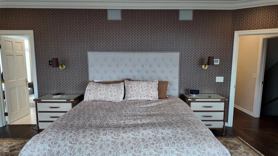 A bedroom with a large bed, white upholstered headboard, two nightstands, two wall-mounted lamps, and patterned wallpaper.