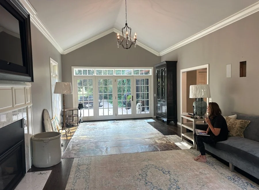 Living room with large glass doors, gray walls, white trim, dark hardwood flooring, a chandelier, a woman sitting on a gray sofa with a laptop, lamps, a black cabinet, and a multicolored rug.