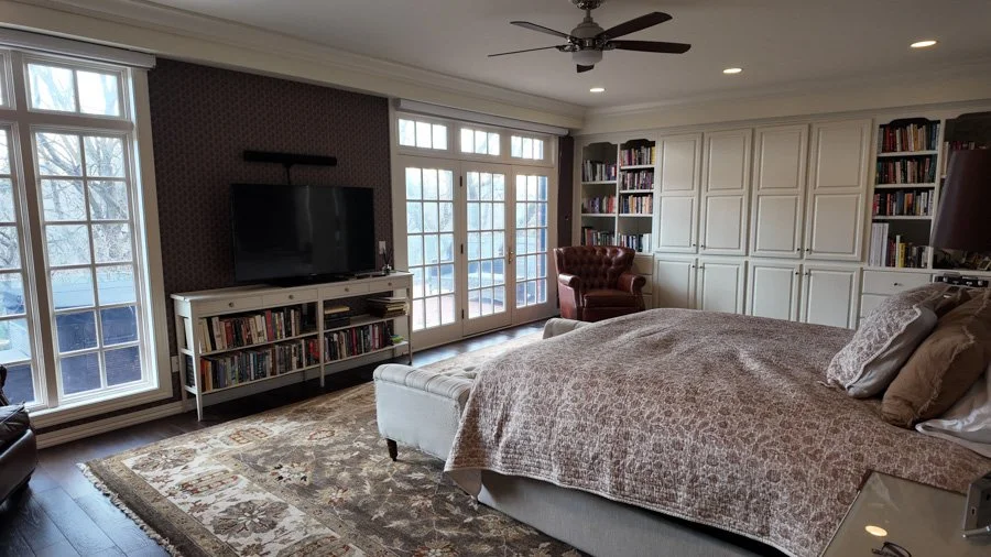 A bedroom with large windows and a glass door, a bed with pillows, a white television stand with a flat-screen TV, a bookshelf against the walls, a reading chair, a ceiling fan, and a patterned area rug.